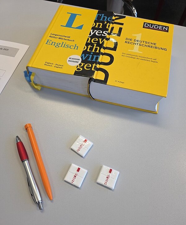 Auf einem Tisch liegen ein Kugelschreiber, ein Bleistift, Traubenzucker und ein englisches und ein deutsches Wörterbuch.