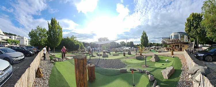 Ein Panoramabild eines Minigolfplatzes mit mehreren Spielern, die auf verschiedenen Bahnen spielen. Der Platz ist von einem Parkplatz umgeben, auf dem mehrere Autos geparkt sind. Im Hintergrund sieht man Bäume, ein Gebäude mit Terrasse und einen sonnigen Himmel mit einigen Wolken. Der Platz ist mit künstlichem Rasen, Steinen und Baumstämmen als Hindernisse gestaltet. Einige Spieler und Spielerinnen sind im Vordergrund und Hintergrund zu sehen.
