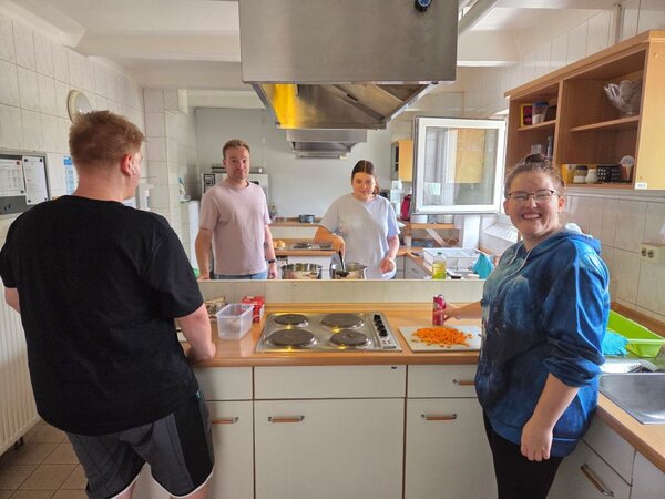 Vier Personen befinden sich in einer Schulküche. Zwei Personen stehen im Vordergrund, eine davon hält ein Schneidebrett mit geschnittenen Karotten. Im Hintergrund sind zwei weitere Personen zu sehen, die ebenfalls in der Küche arbeiten. Die Küche ist mit verschiedenen Küchengeräten und Utensilien ausgestattet, darunter ein Herd, ein Kühlschrank, und mehrere Schränke. Es gibt auch verschiedene Küchenutensilien auf der Arbeitsplatte.