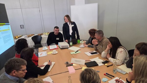 Eine Frau mit Namensschild steht vor einer Gruppe von Personen, die um einen großen Tisch in einem Seminarraum sitzen. Die Teilnehmenden hören aufmerksam zu und machen sich Notizen. Auf dem Tisch liegen Unterlagen, Stifte und Getränke. Ein Beamer und ein Laptop sind ebenfalls zu sehen.