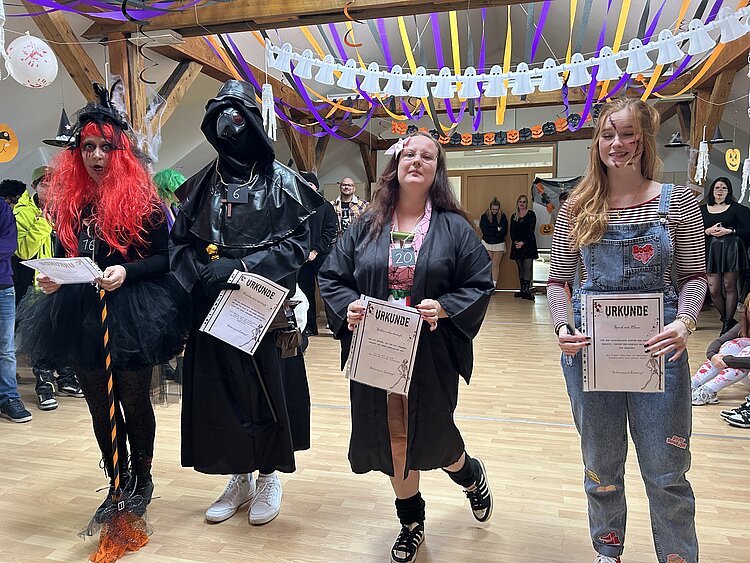 Vier kostümierte Teilnehmende halten Urkunden in einem festlich mit Geister- und Kürbisgirlanden geschmückten Saal – Kostümwettbewerb.