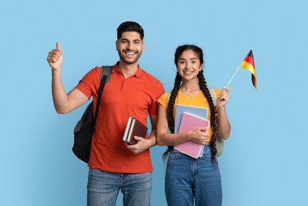 Eine junge Frau mit arabischem Aussehen schwenkt eine Deutschlandflagge, ein junger Mann mit südländischem Teint macht ein "Daumen hoch"-Zeichen. Beide haben Bücher in der Hand und Rücksäcke auf.
