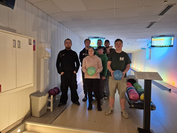 Gruppenfoto auf der Bowlingbahn: Mehrere Teilnehmende stehen gemeinsam am Bahnanfang, zwei halten Bowlingkugeln in den Händen; im Hintergrund sind Anzeigetafeln und Kugelrücklauf zu sehen.