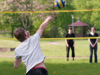 Ein junger Mann in einem weißen T-Shirt und dunklen Shorts spielt Volleyball im Freien. Er schlägt einen gelb-blauen Volleyball über ein gelbes Netz. Im Hintergrund stehen zwei Personen und beobachten das Spiel. Die Szene spielt auf einem grünen Rasen mit Bäumen und einem Pavillon im Hintergrund.