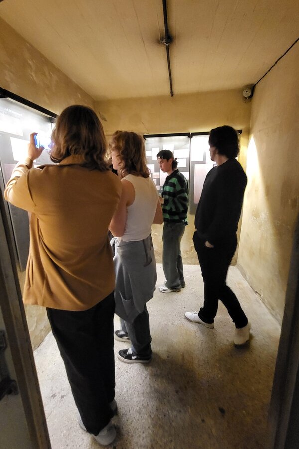 Vier Teilnehmende des Jugendintegrationskurses stehen in einem schmalen, gelblich beleuchteten Flur mit rauen Wänden. Eine Person im Vordergrund hält ein Smartphone und fotografiert oder filmt durch ein Fenster auf der linken Seite. Die anderen Personen stehen nebeneinander und blicken ebenfalls in Richtung des Fensters. Alle tragen legere Kleidung und helle Schuhe. Im Hintergrund sind weitere Fenster mit undurchsichtigen Scheiben zu sehen.