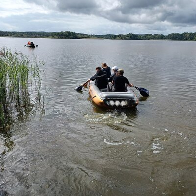 Auf einem See sind zwei Menschengruppen in Schlauchbooten mit Paddeln unterwegs. Vorne links ist Schilf zu sehen. Im Hintergrund ist ein bewaldetes Seeufer zu sehen.
