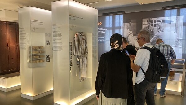 Teilnehmende stehen in der Gedenkstätte Sachsenhausen und betrachten Exponate, darunter gestreifte KZ-Häftlingskleidung. 