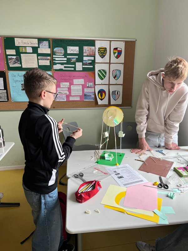 Zwei Jungen stehen an einem Tisch und basteln ein Modell mit Papier, Zahnstochern und weiteren Bastelmaterialien. Auf dem Tisch liegen bunte Papiere, Scheren und Anleitungen.