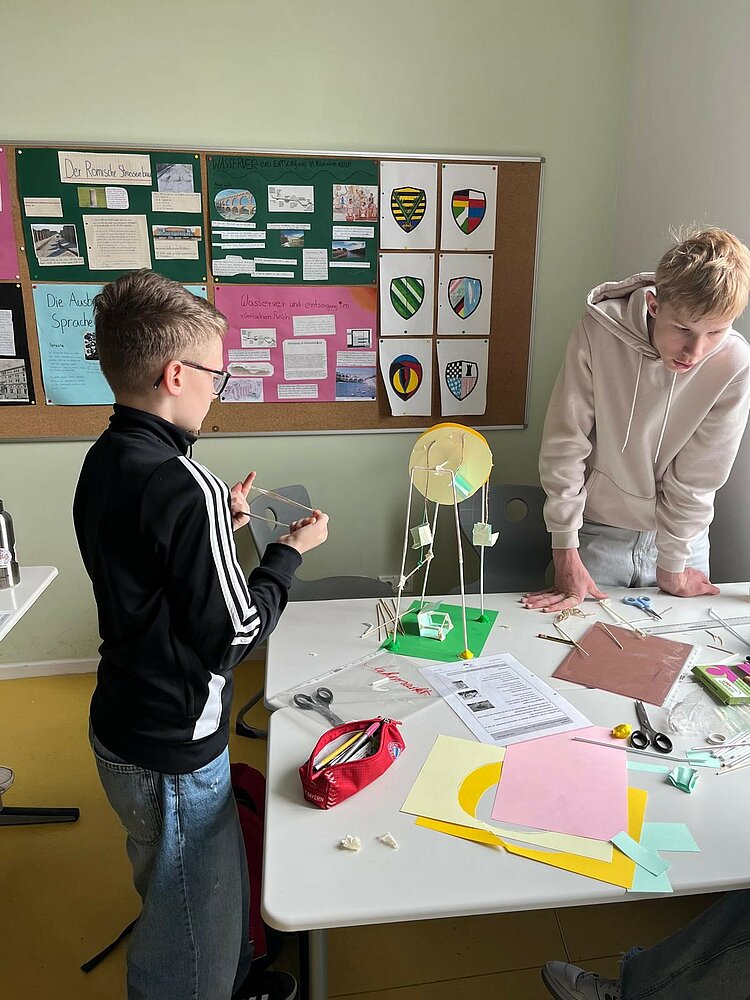 „Praxisberater*innen an Oberschulen“ – Potentialanalyse Zwei Jungen stehen an einem Tisch und basteln ein Modell mit Papier, Zahnstochern und weiteren Bastelmaterialien. Auf dem Tisch liegen bunte Papiere, Scheren und Anleitungen.