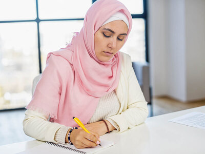 Eine Frau mit einem rosa Kopftuch und einem beigen Pullover sitzt an einem Tisch und schreibt in ein Notizbuch. Sie hält einen gelben Stift in der Hand. Der Hintergrund zeigt ein helles, modernes Büro oder Klassenzimmer mit großen Fenstern.