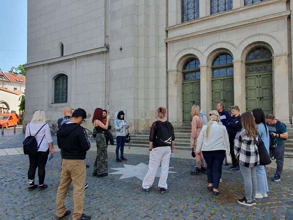  Eine Gruppe von Menschen steht vor einem historischen Gebäude mit großen, grünen Doppeltüren und hohen Fenstern. Die Gruppe scheint sich auf eine Führung oder Besichtigung vorzubereiten.