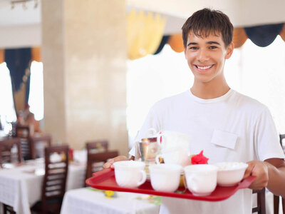 Ein junger Mann mit einem freundlichen Lächeln und weißem T-Shirt steht in einem Restaurant oder Hotel. Er trägt ein rotes Tablett mit weißem Geschirr, darunter Tassen, eine kleine Kaffeekanne und eine Zuckerstreudose. Im Hintergrund sind gedeckte Tische und ein Buffetbereich zu sehen. Der Raum wirkt hell und einladend.