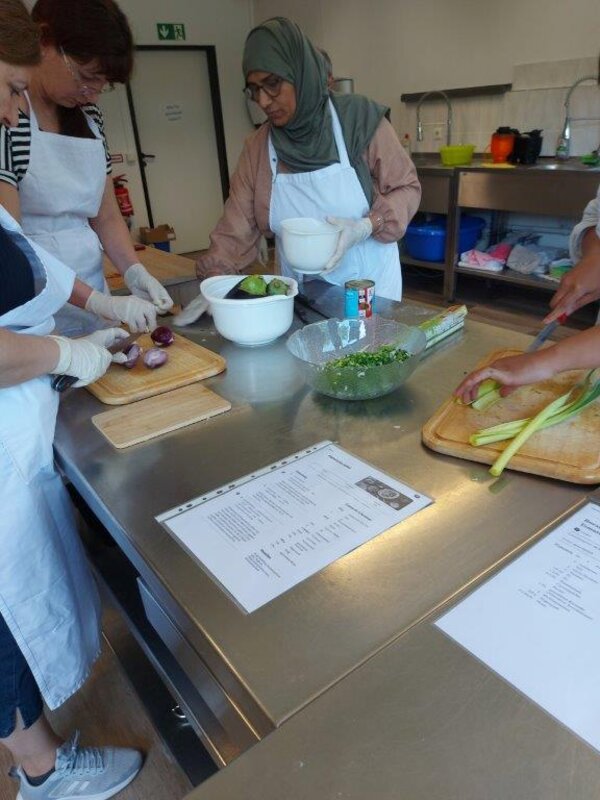 Mehrere Personen mit Schürzen schneiden Gemüse auf einem Edelstahltisch, darunter rote Zwiebeln und Lauch. Auf dem Tisch liegen zudem Rezeptblätter und Schüsseln mit vorbereiteten Zutaten.