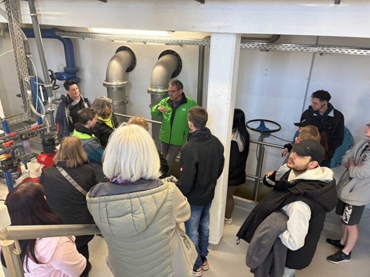 Teilnehmende Auszubildende in den Wasserwerken Hoyerswerda stehen um einen Mitarbeiter herum, in einer industriellen Halle mit großen Rohren und hören ihm zu.