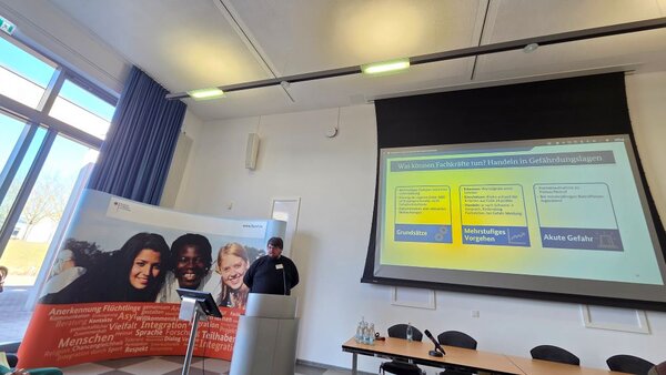 Ein Referent hält einen Vortrag in einem Seminarraum. Auf der Leinwand hinter ihm ist eine Präsentation mit dem Titel ‚Was können Fachkräfte tun? Handeln in Gefährdungslagen‘ zu sehen. Die Folie zeigt drei Handlungsmöglichkeiten: Grundsätze, Mehrstufiges Vorgehen und Akute Gefahr. Im Raum sind Stühle und ein Tisch mit Informationsmaterialien zu erkennen.