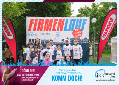 Gruppenfoto von Teilnehmenden beim Firmenlauf vor einer großen Veranstaltungswand mit der Aufschrift „Firmenlauf“; mehrere Personen tragen Laufshirts und Startnummern.
