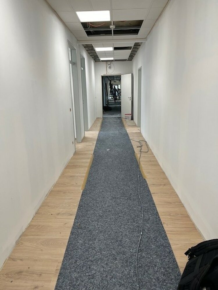 Langer heller Flur mit neu verlegtem Holzfußboden und einem grauen Schutzteppich in der Mitte. An der Decke sind einige Platten noch offen, Kabel hängen herab – der Bereich befindet sich sichtbar in Renovierung.