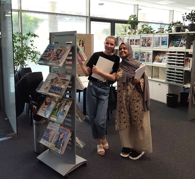 Gruppe IK-359 zu Besuch im BIZ Zwei Teilnehmende des Integrationskurses stehen lächelnd vor einem Informationsständer im Berufsinformationszentrum und halten Broschüren in den Händen.