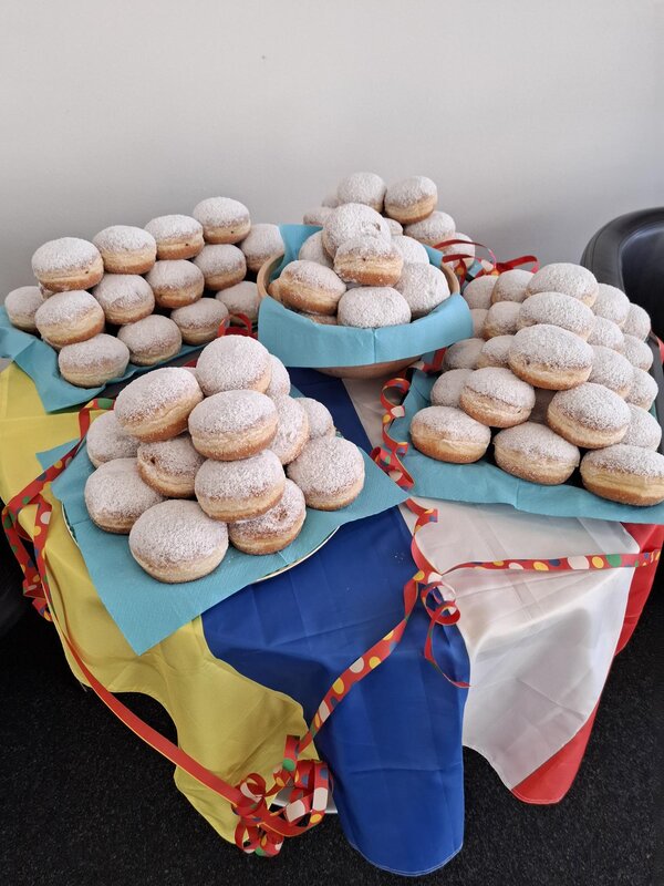 Auf einem Tisch mit eine blau, gelb, rot und weißen Fahne liegen viele Berliner mit Puderzucker auf Tellern gestapelt.