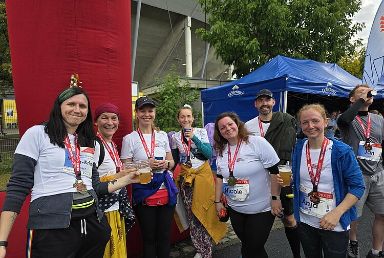 Euro-Schulen & Euro Akademie bei der Rewe Team Challenge 2025 Gruppe von neun Teilnehmenden steht mit Medaillen und Getränken vor dem Zielbereich. Alle lachen oder stoßen an. Im Hintergrund ist ein blauer Lichtenauer-Zeltpavillon zu sehen.