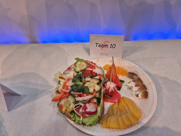 Ein kunstvoll belegtes Brot mit Feldsalat, Gurke, Radieschen, Paprika und Salat, daneben Pfirsichscheiben und Mandarinen auf einem Pappteller; dahinter ein Schildchen mit der Aufschrift "Team 10".