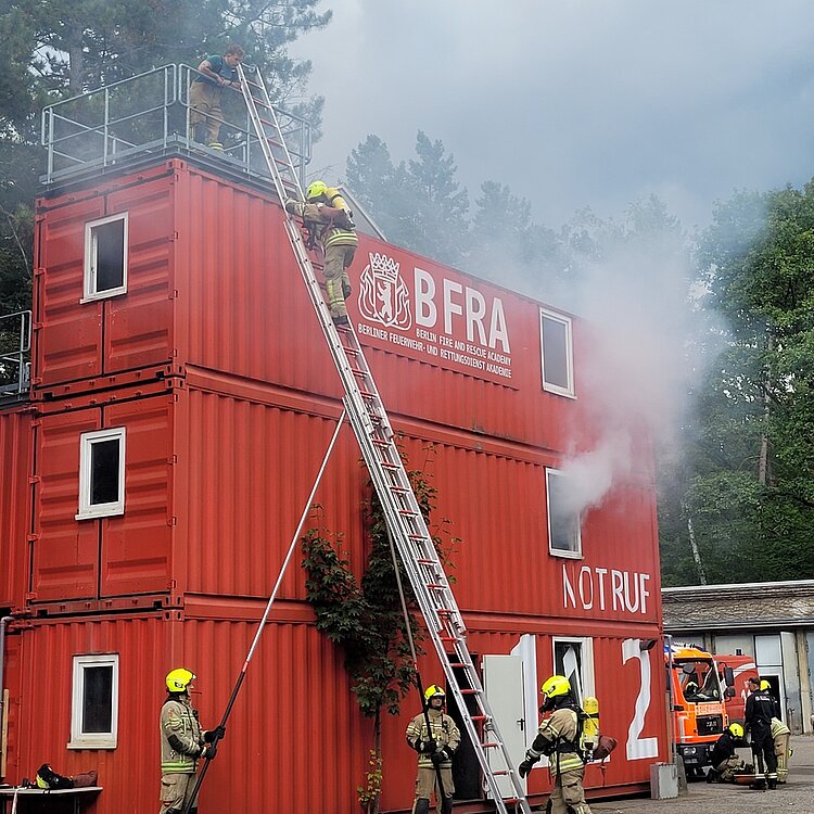 Die Feuerwehr bei einer Übung: Drei rote Wohncontainer stehen aufeinander, aus einem Zimmer dringt dichter Rauch. Zwei Feuerwehrmänner sichern die Leiter, die auf das Dach der Container führt. Eine Person in Ausrüstung mit Sauerstoffflasche auf dem Rücken hat fast das Ende der Leiter erreicht und wird oben von einem Kollegen erwartet, eine zweite Person ist im Begriff, die Leiter zu besteigen.