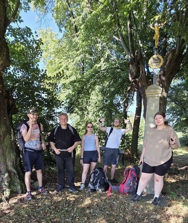 Fünf Personen stehen lächelnd in einer Gruppe unter Bäumen mit dichtem grünem Laub. Im Hintergrund ist ein christliches Kreuz mit einer goldenen Jesusfigur auf einem Sockel zu erkennen.
