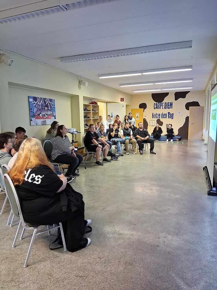 Eine Gruppe Jugendlicher sitzt in einem Klassenraum auf Stühlen und schaut auf eine Leinwand, auf der das „KifferQuiz“ projiziert wird. Im Hintergrund steht an der Wand der Schriftzug „Carpe Diem – Nutze den Tag“.