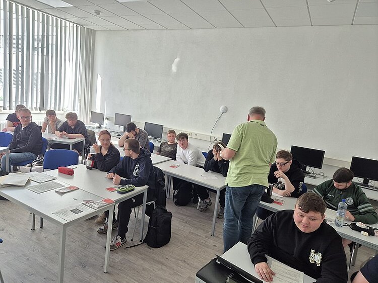 Ein Lehrer in grünem Hemd verteilt Unterlagen an Schüler, die an Gruppentischen sitzen. Im Raum stehen Computer, viele Schüler schauen auf ihre Blätter oder lachen. Ein Schüler vorne rechts lächelt direkt in die Kamera.