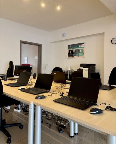 Ein moderner Schulungs- oder Seminarraum mit mehreren Arbeitsplätzen. Auf langen, hellen Tischen stehen Laptops, Mäuse und Kabel bereit. Jeder Platz ist mit einem Bürostuhl ausgestattet. Im Hintergrund sind ein Drucker, ein Schrank und ein Wandbild mit der Aufschrift 'EVER' zu sehen. Der Raum wirkt aufgeräumt und gut organisiert.