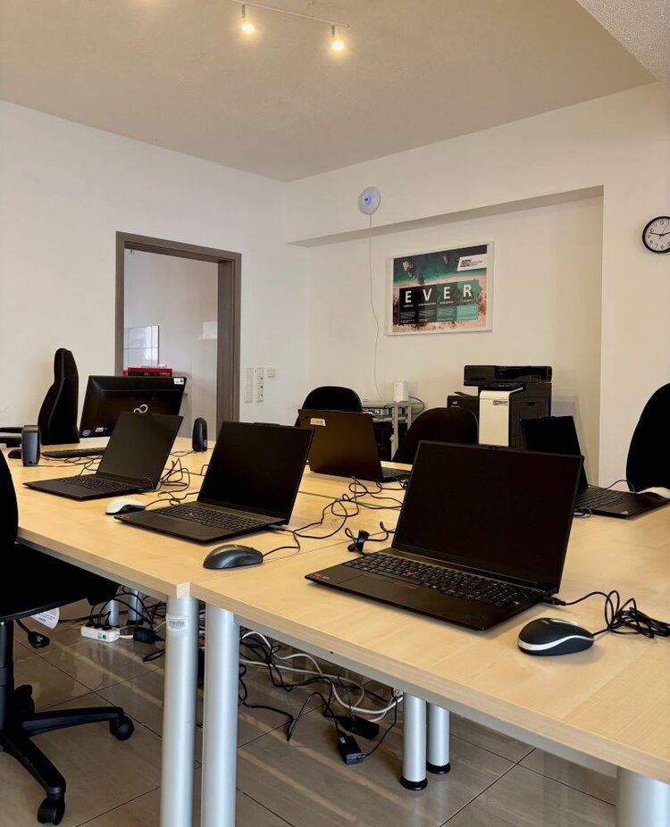 Ein moderner Schulungs- oder Seminarraum mit mehreren Arbeitsplätzen. Auf langen, hellen Tischen stehen Laptops, Mäuse und Kabel bereit. Jeder Platz ist mit einem Bürostuhl ausgestattet. Im Hintergrund sind ein Drucker, ein Schrank und ein Wandbild mit der Aufschrift 'EVER' zu sehen. Der Raum wirkt aufgeräumt und gut organisiert.