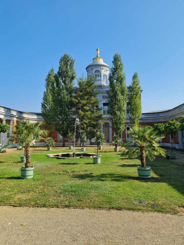 Ein mit Palmen und Baumen begrünter Bereich mit Springbrunnen. Im Hintergrund ist ein Teil eines Schlosses zu sehen.