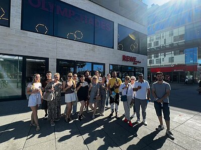 Heiße Köpfe, kühles Eis – Sprachkurse bei 35 Grad! Eine Gruppe von etwa 18 jungen Erwachsenen steht bei sonnigem Wetter vor dem Einkaufszentrum Olymp & Hades in Leipzig. Im Hintergrund sind Schilder von REWE und Vodafone zu sehen. Die Gruppe hält Eis in der Hand und lächelt in die Kamera.