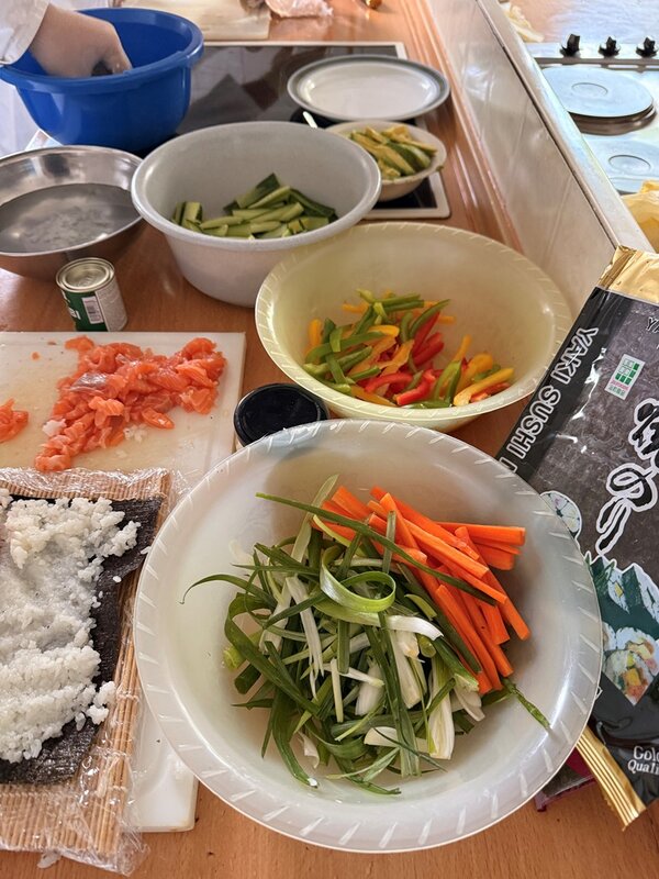 Im Vordergrund stehen mehrere Schüsseln mit geschnittenem Gemüse, darunter Karotten, Frühlingszwiebeln, Gurken und Paprika. Links liegt eine Portion roher Lachs auf einem Schneidebrett. Daneben befindet sich eine Bambusmatte mit Reis, die für Sushi verwendet wird. Rechts ist eine Verpackung mit der Aufschrift Sushi Nori zu sehen. Im Hintergrund arbeitet eine Person mit einer blauen Schüssel. Die Szene zeigt die Vorbereitung von Sushi-Zutaten auf einer Küchenarbeitsfläche.