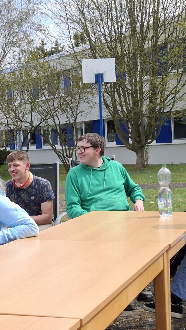 Zwei junge Männer sitzen lächelnd an einem langen Tisch im Freien. Einer trägt ein grünes Sweatshirt, der andere ein dunkles T-Shirt. Auf dem Tisch steht eine Flasche Wasser.