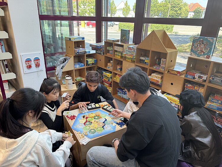 Lernorte erkunden – ein Ausflug in die Stadtbibliothek Wolfen Eine Gruppe Jugendlicher und Kinder sitzt an einem Tisch und spielt das Brettspiel „Die Siedler von Catan“. Im Hintergrund stehen Regale voller Gesellschaftsspiele. Der Raum ist hell mit großen Fenstern, durch die man nach draußen sieht.