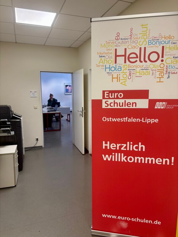 In einem weißen Flur steht rechts ein großes Roll-Up der Euro-Schulen mit der Aufschrift "Herzlich Willkommen!"; dahinter ist eine offene Tür zu einem Klassenzimmer mit einer Person zu sehen.