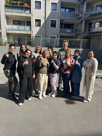 Eine Gruppe von Erwachsenen steht bei sonnigem Wetter in einem Innenhof vor einem Wohngebäude. Mehrere Personen zeigen mit erhobenem Daumen in die Kamera und posieren gemeinsam für ein Gruppenfoto.