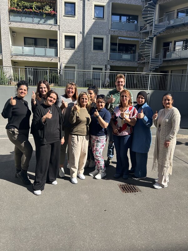 Eine Gruppe von Erwachsenen steht bei sonnigem Wetter in einem Innenhof vor einem Wohngebäude. Mehrere Personen zeigen mit erhobenem Daumen in die Kamera und posieren gemeinsam für ein Gruppenfoto.