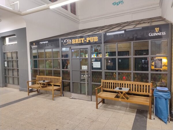 Außenansicht des „The Brit-Pub“ in einem Flur mit Fliesenboden; vor der Glasfront mit Logos von Guinness stehen zwei Holzbänke mit kleinen Tischen.