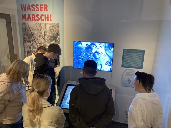 Eine Gruppe von Menschen steht vor einer interaktiven Ausstellung mit dem Thema Wasser. Sie schauen auf einen Bildschirm mit einer Nahaufnahme von Wassertropfen. Im Hintergrund ist ein Plakat mit der Aufschrift „WASSER MARSCH!“ zu sehen.
