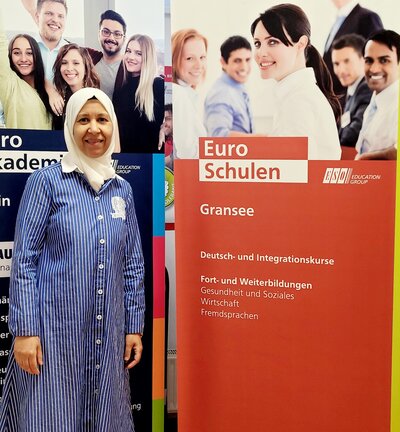 Eine Frau mit einem langen blau-weißen Kleid und einen Kopftuch steht neben dem Roll-up der Euro-Schulen.