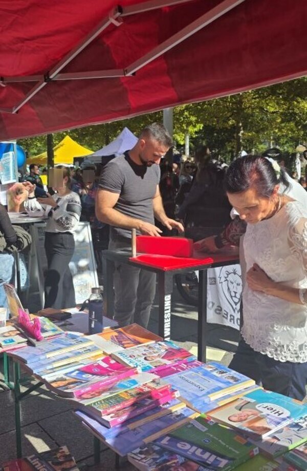 Eine Besucherin mit einem weißem Oberteil aus Spitze informieren sich an einem Infostand unter rotem Zelt. Der Tisch ist mit bunten Büchern und Broschüren gefüllt. Im Hintergrund sind weitere Personen zu sehen.
