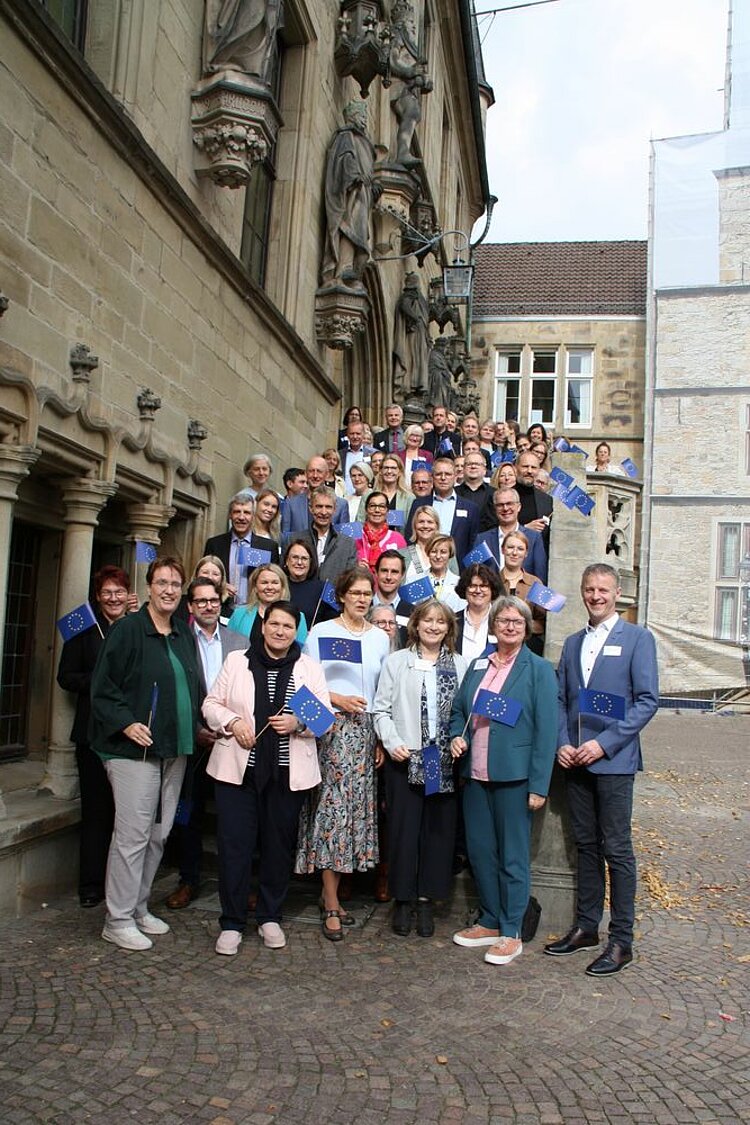 Euro Akademie Oldenburg: weiterhin „die“ Europaschule in Niedersachsen Die Schulleitungen der "Europaschulen in Niedersachsen" haben sich für ein Gruppenfoto auf der Treppe des Osnabrücker Rathauses aufgestellt. Sie halten alle kleine Europaflaggen aus Papier in der Hand.