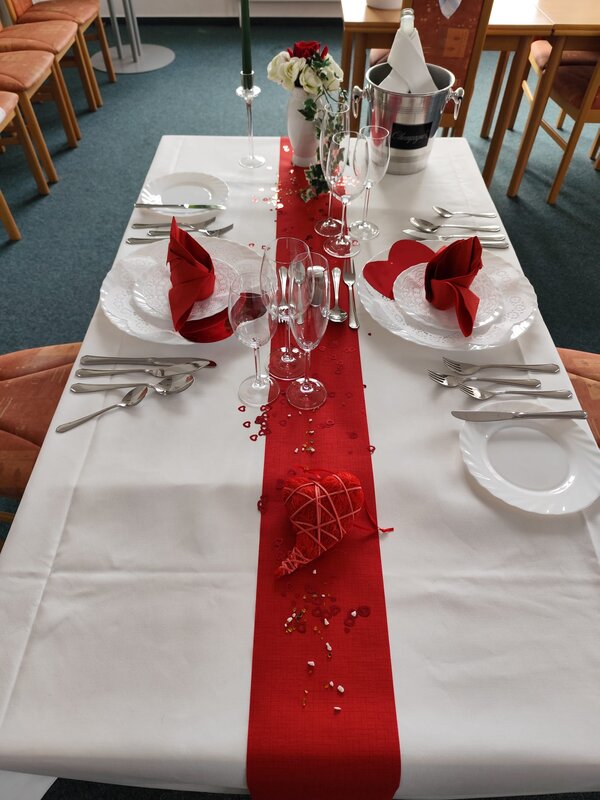 Eine stilvolle Tischdekoration mit einem roten Läufer, roten Servietten und transparenten Weingläsern. In der Mitte liegt ein dekoratives, geflochtenes rotes Herz.