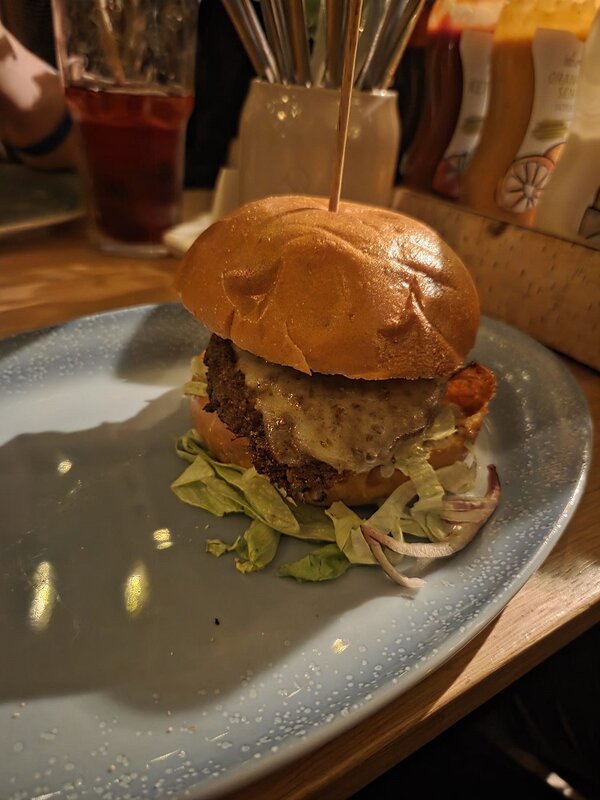Ein Burger mit hellem Brötchen, gebratenem Patty und geschmolzenem Käse liegt auf einem hellblauen Teller. Unter dem Burger befinden sich einige Salatblätter und rote Zwiebelringe. Im Hintergrund stehen Gläser, Flaschen und Saucenflaschen auf einem Holztresen. Die Szene ist warm beleuchtet.