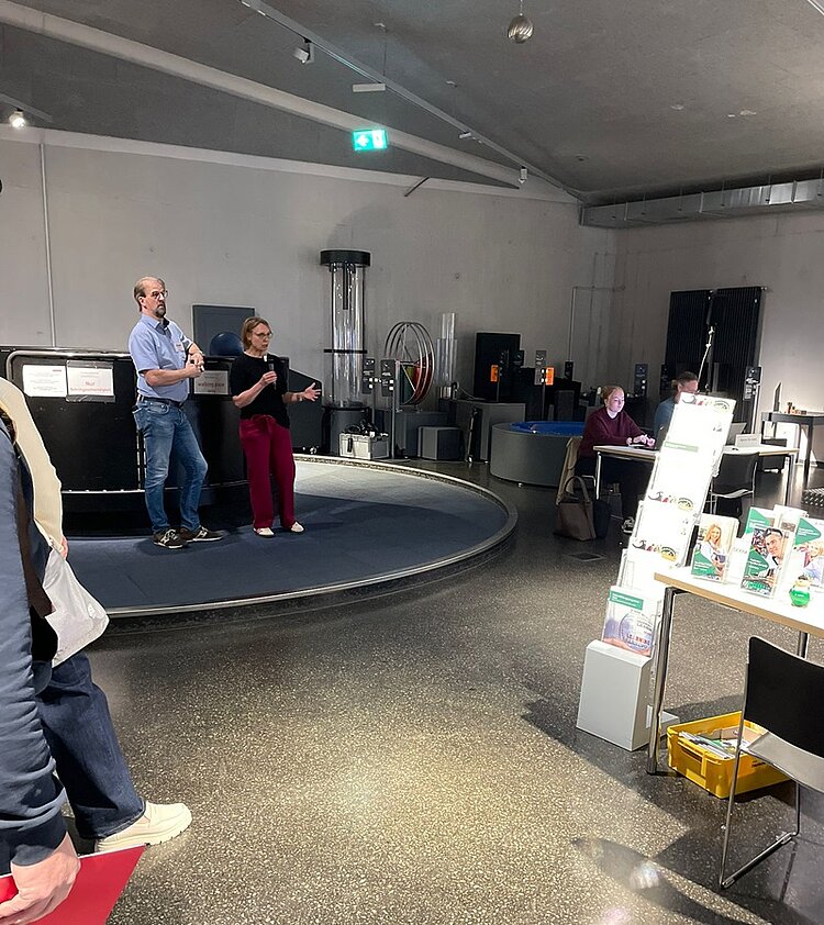 Erfolgreiches Bildungs-Speed-Dating Zwei Moderator*innen führen durch die Veranstaltung Bildungs-Speed-Dating in der Phänomenta in Lüdenscheid. Sie stehen auf der Bühne, vor ihnen sind Informationsstände der Teilnehmenden zu erkennen, mit Broschüren und Katalogen.