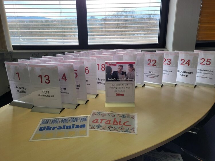 Tisch mit nummerierten Aufstellern für die Arbeitgeber beim Job-Speed-Dating der Euro-Schulen Märkischer Kreis in der Phänomenta in Lüdenscheid: In mehreren Reihen stehen transparente Aufsteller mit gut lesbaren roten Nummern und den Namen teilnehmender Unternehmen. In der Mitte befindet sich ein Schild mit der Aufschrift ‚Job-Speed-Dating‘, inklusive Hinweis auf gute Gespräche und Erfolg. Vor dem Tisch liegen dekorative Ausdrucke mit den Schriftzügen ‚Ukrainian‘ und ‚Arabic‘, die auf vorhandene sprachliche Unterstützung hinweisen. Im Hintergrund ist ein großes Fenster mit Blick auf eine leicht hügelige Landschaft.