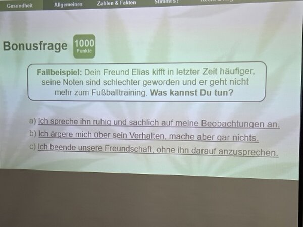 Eine Quizfrage auf der Leinwand thematisiert, wie man reagieren kann, wenn ein Freund zunehmend Cannabis konsumiert und sich sein Verhalten verändert.