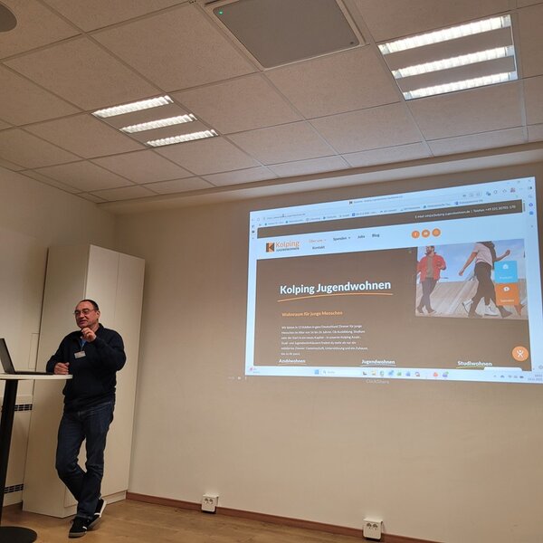 Ein Mann steht in einem Seminarraum neben einem Stehtisch und spricht vor einer projizierten Präsentationsfolie zum Thema ‚Kolping Jugendwohnen‘. Auf der Leinwand ist die Webseite des Kolping Jugendwohnens gut erkennbar dargestellt. Der Raum ist modern eingerichtet, mit heller Wandfarbe, Holzfußboden und einer abgehängten Decke mit LED-Paneelen. Das Bild entstand während des 21. Reinickendorfer Ausbildungsdialogs an den Euro-Schulen Berlin.